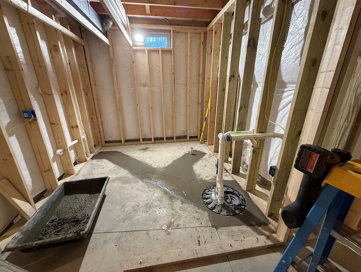 An unfinished basement room with wood wall framing, a concrete floor with fresh patches, and exposed plumbing pipes.