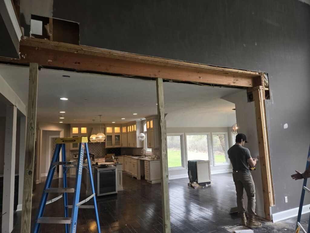A person works on a wooden support beam framing a wide doorway in a house under renovation, with a kitchen visible behind.