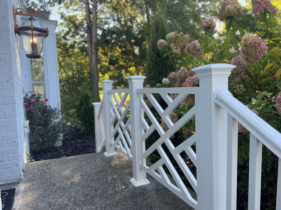 A white lattice-style porch railing stands on a stone walkway next to a light fixture and blooming hydrangea bushes.