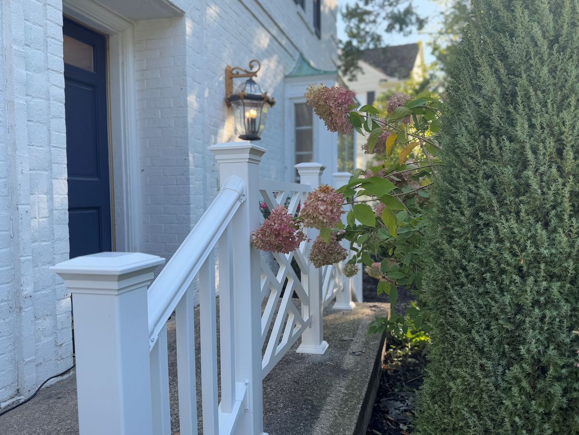 A navy blue front door with white brick walls, a white lattice railing, a wall-mounted lantern, and pink hydrangeas.