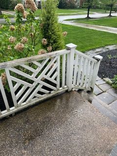 White wooden railing with a geometric, crisscross design leading down stone stairs beside a blooming bush and lawn.