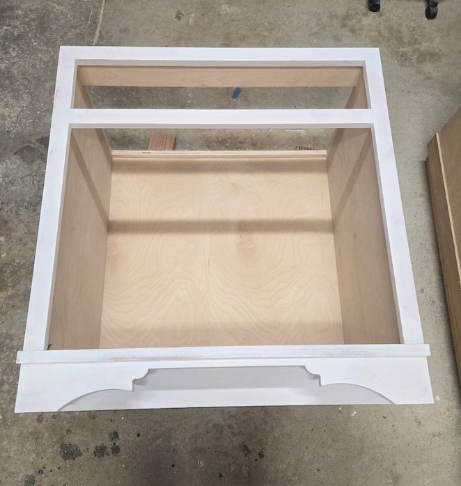 A top-down view of an unfinished white wooden vanity cabinet frame with plywood interior sides and a decorative apron.