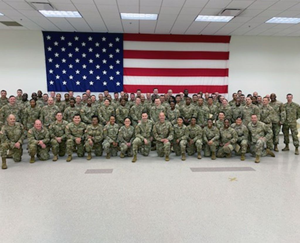 A large group of soldiers in military uniforms standing in rows before a large American flag.