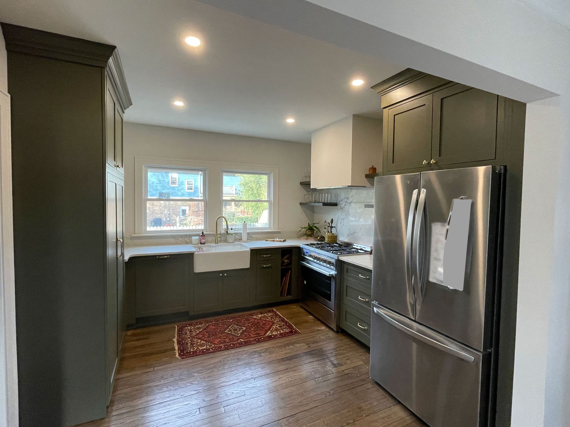 A modern, renovated kitchen featuring dark green cabinetry, a stainless steel refrigerator, and a white farmhouse sink.