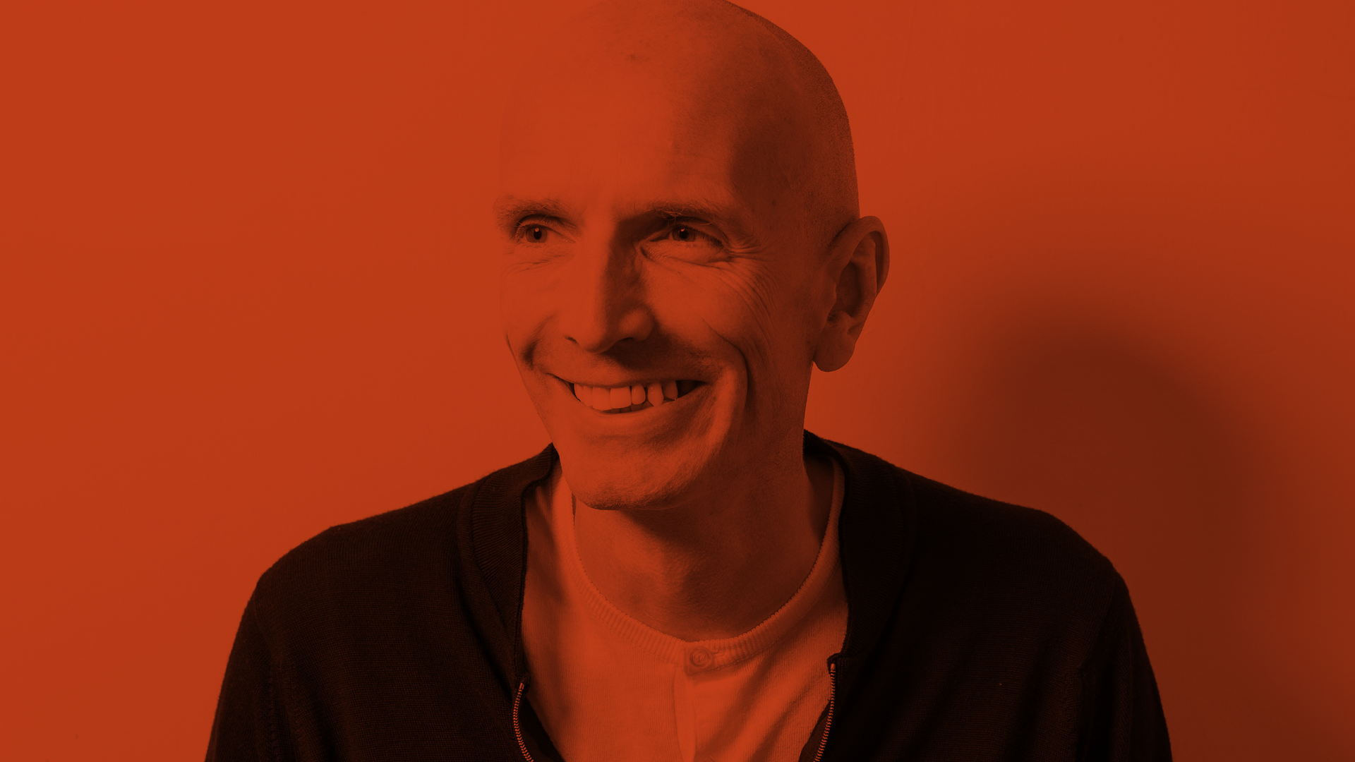 A red and black duotone image of a smiling bald middle-aged man wearing a casual black jacket