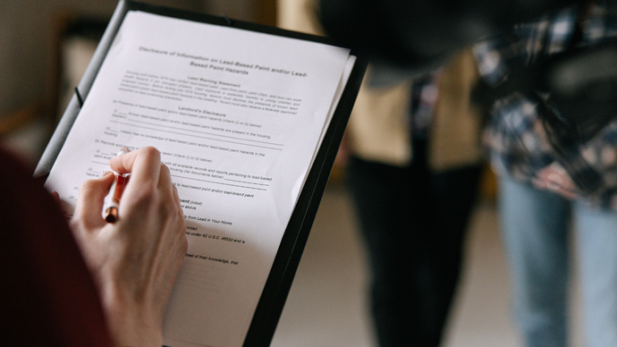 A close-up view of a person writing on a document held on a clipboard, with blurred individuals in the background.