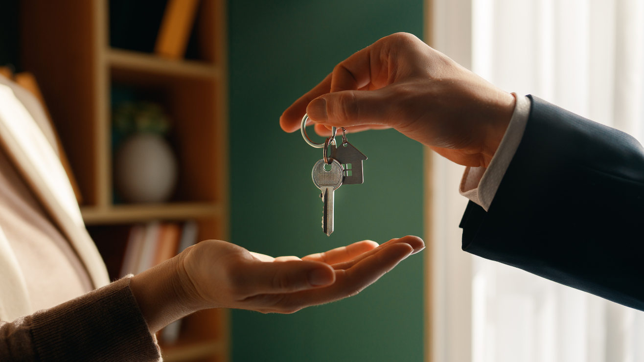 A person in a dark suit hands house keys to an open hand in an indoor setting.