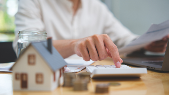 A person uses a calculator next to a small house model and a jar of coins, suggesting home finance planning.
