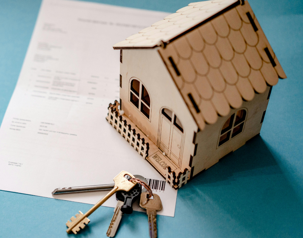 A small wooden model house sits on a document next to a set of metal keys against a teal background.