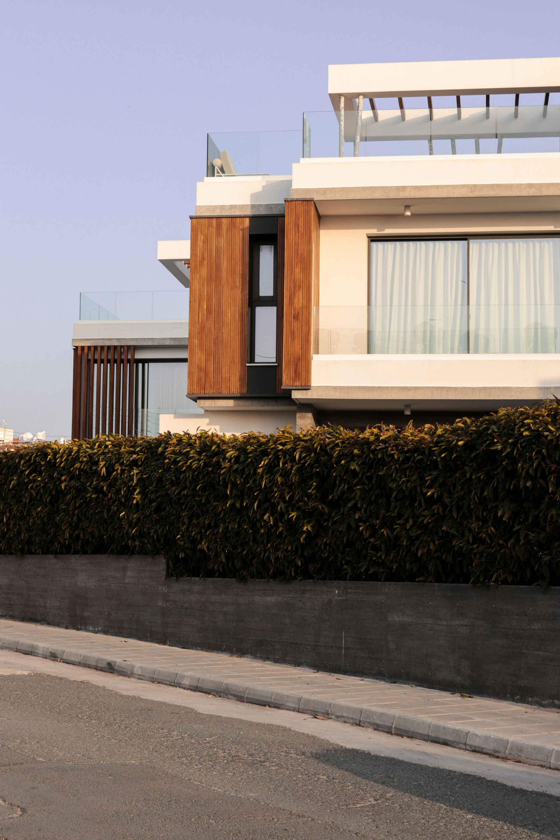 A modern house with white concrete walls, wood paneling, and a balcony, behind a tall, dark hedge and stone wall.