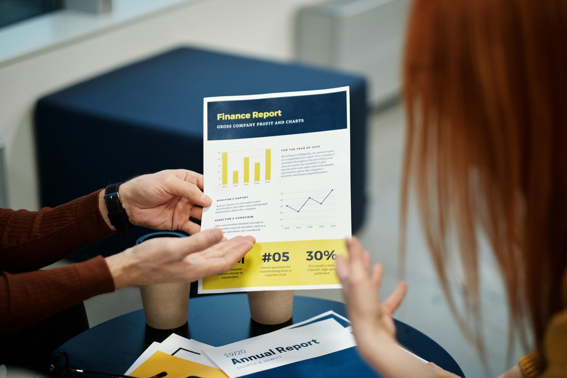 Two people discuss a progress report with charts and data displayed on a printed document in a professional setting.