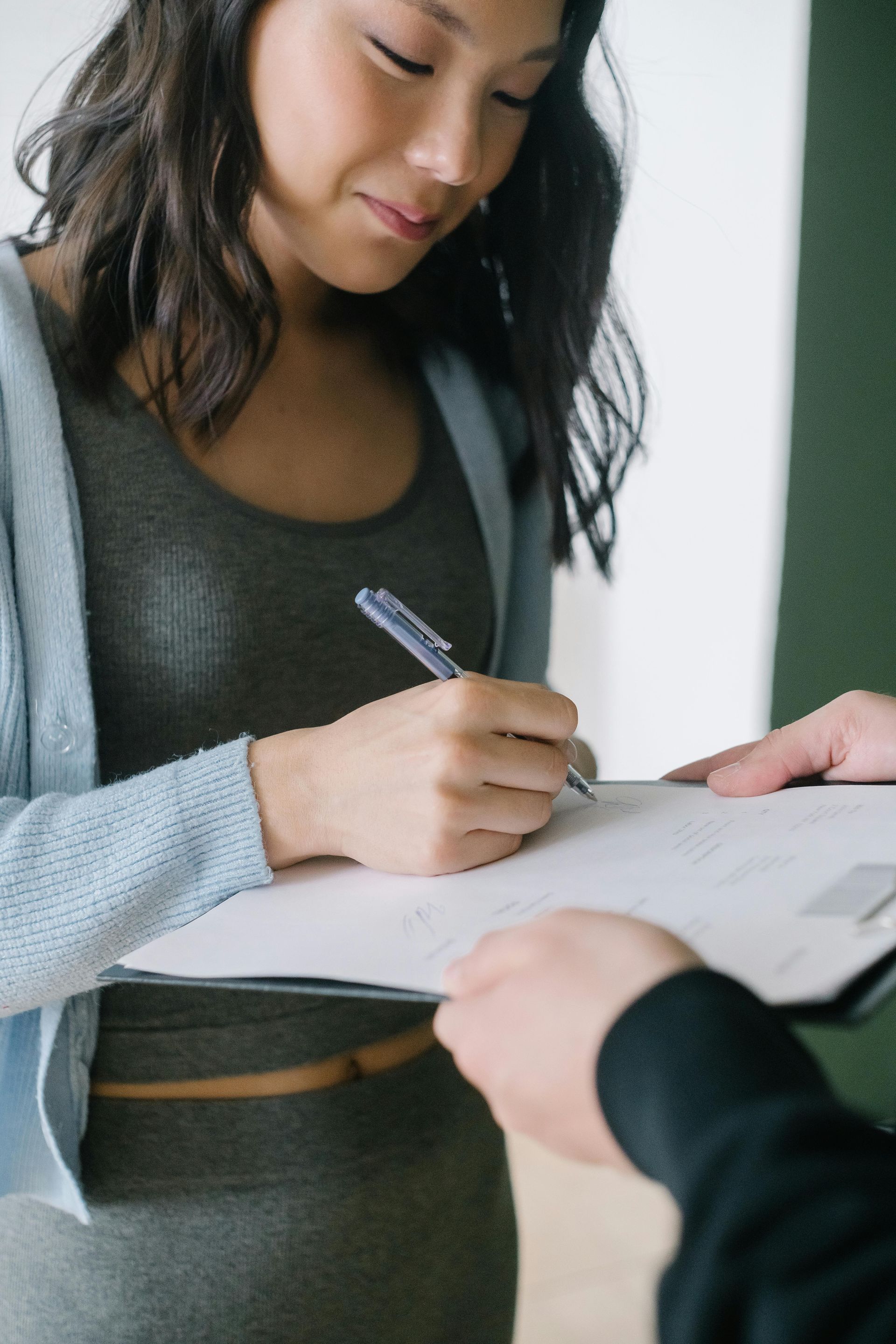 A person wearing a light blue cardigan and grey top writes on a clipboard held by another person.