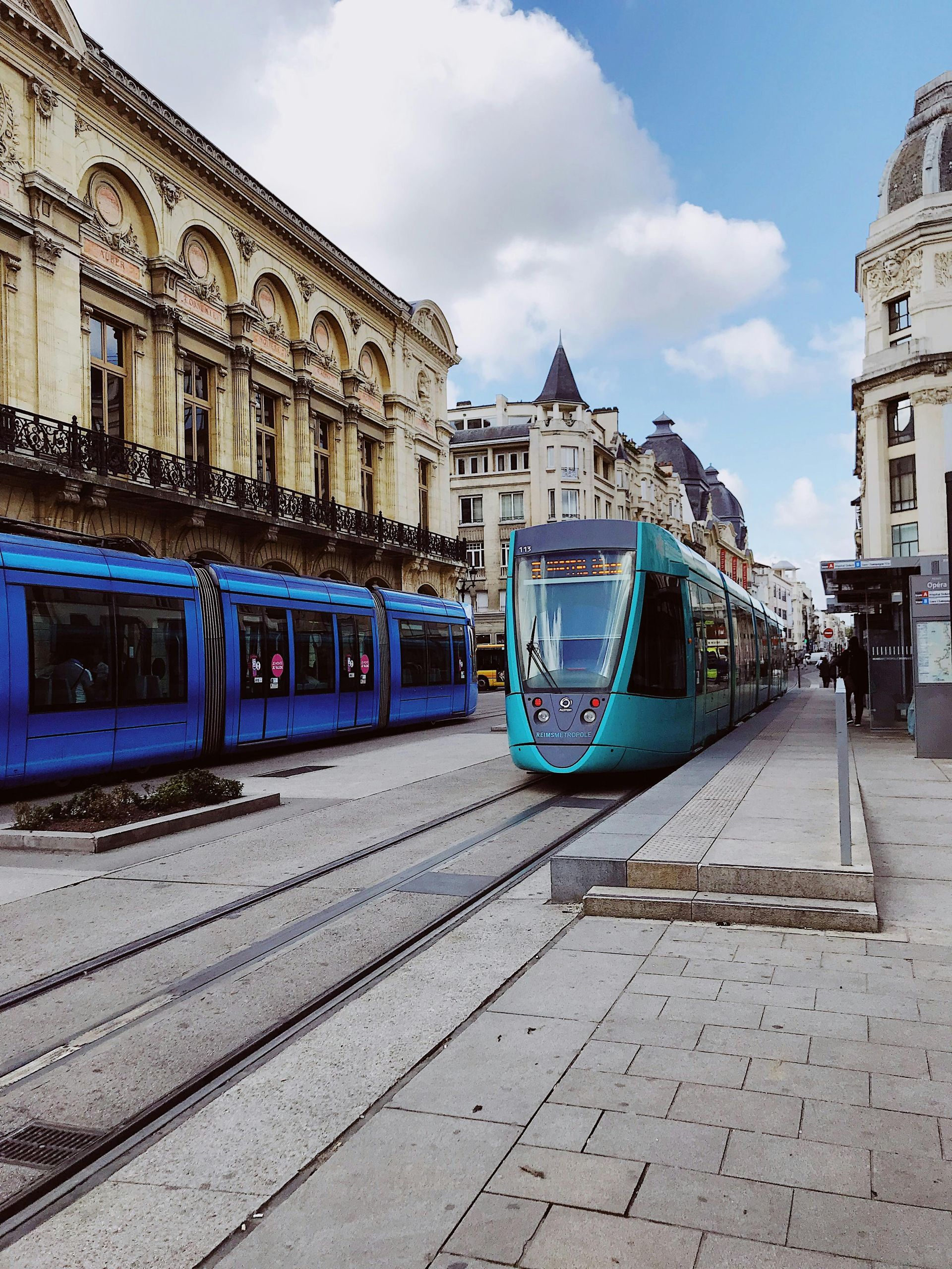 Deux tramways bleus sur les rails d'une ville européenne. De hauts bâtiments ornés bordent la rue, sous un ciel nuageux.