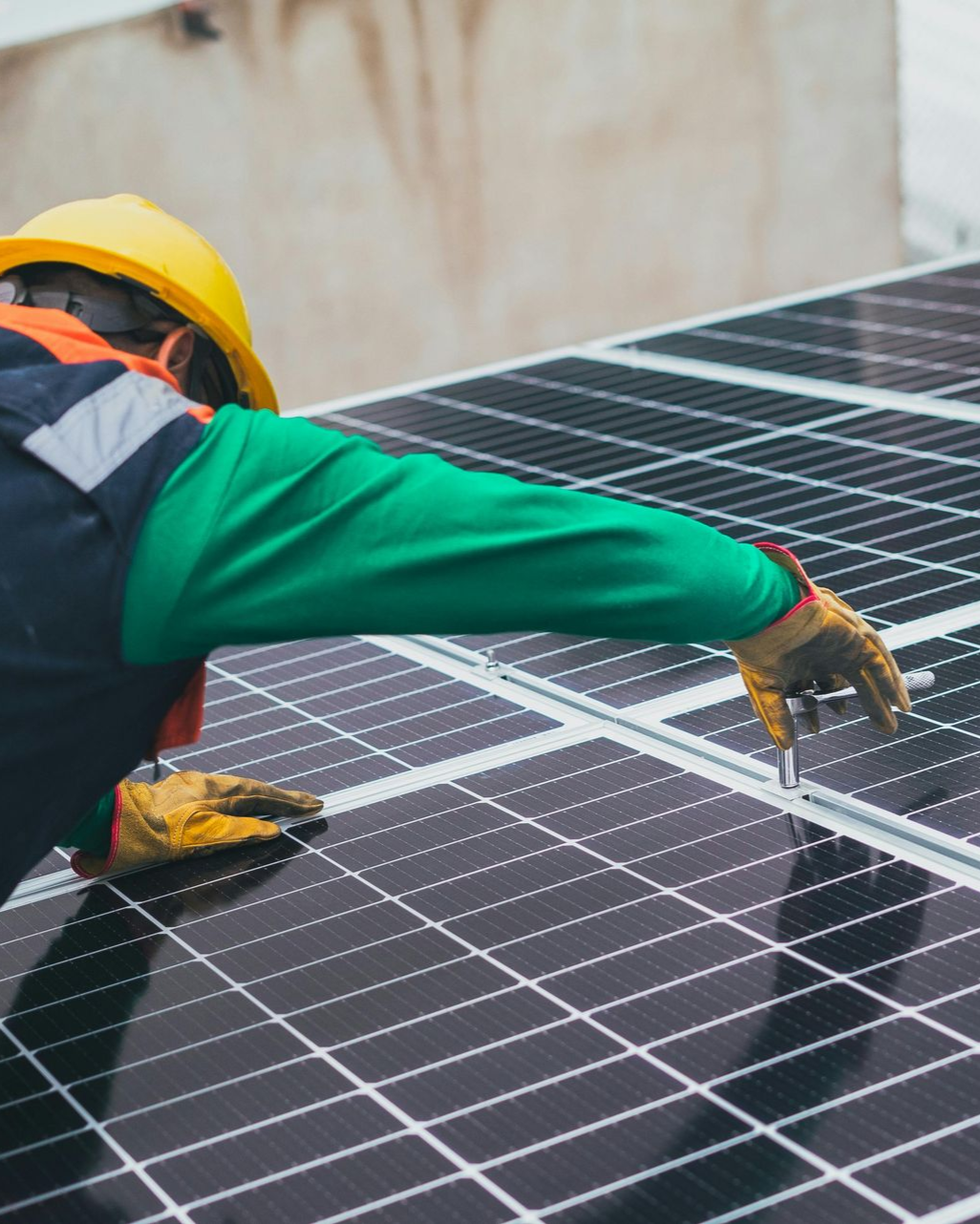 Technicien en panneaux solaires, casque jaune, inspectant un réseau de panneaux solaires. Chemise verte, gants beiges.