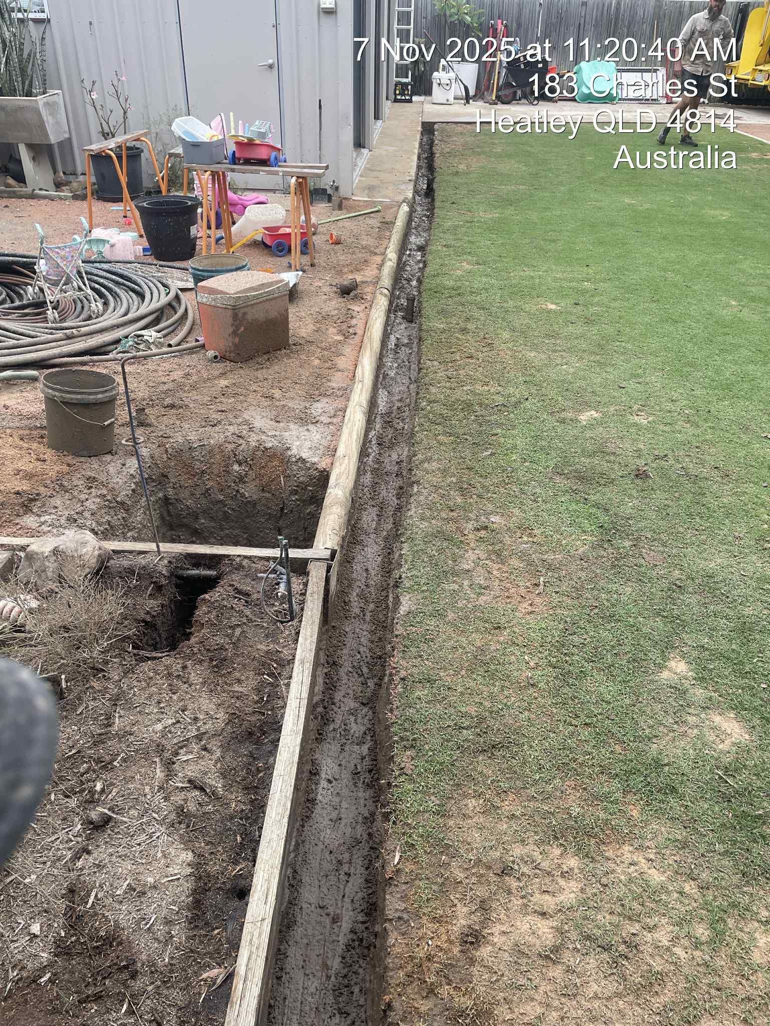 Backyard trench and exposed pipes beside a grassy lawn, with construction tools and materials along the fence line