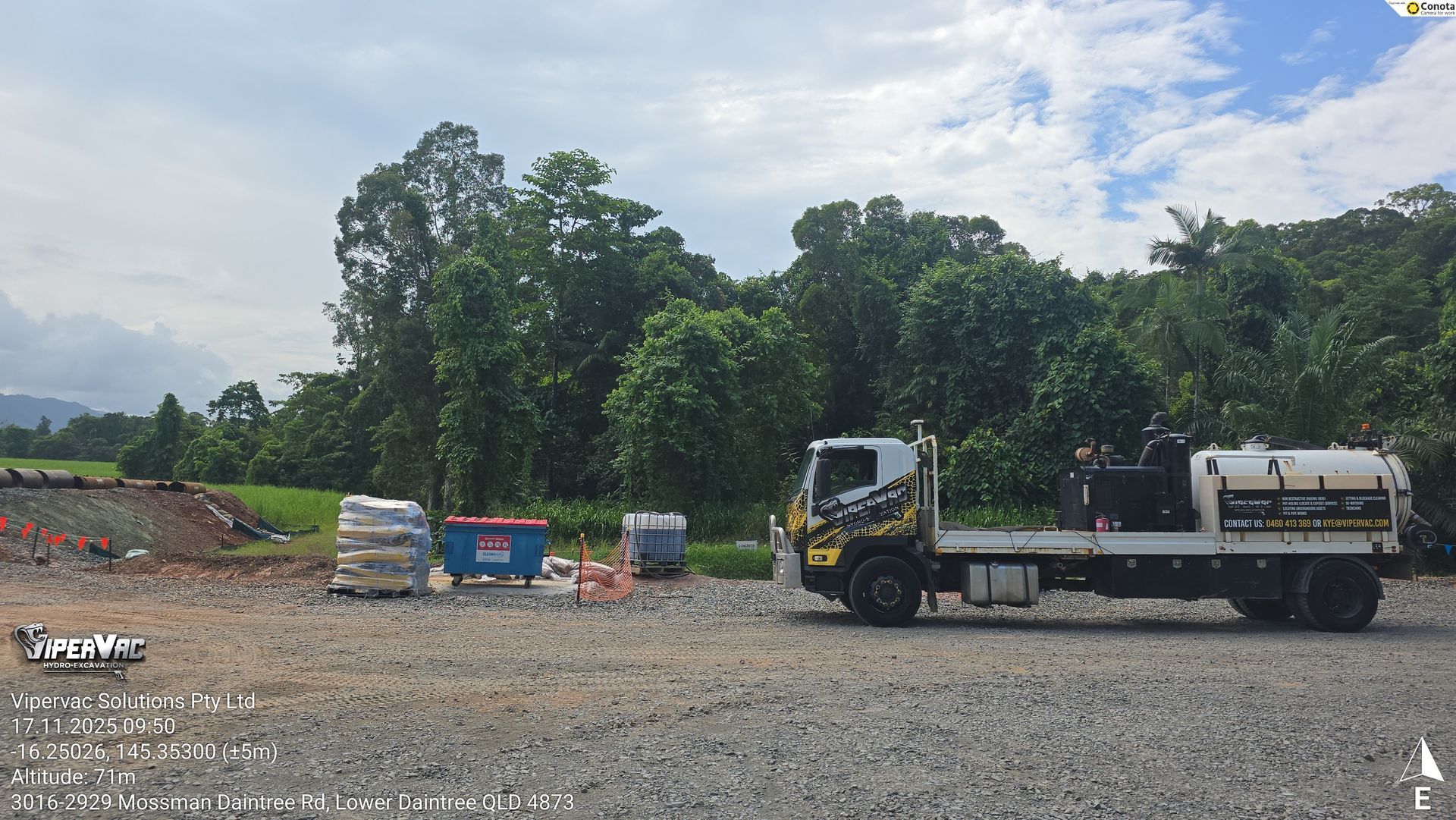 Construction site with gravel lot, white dump truck, and large trees under a partly cloudy sky