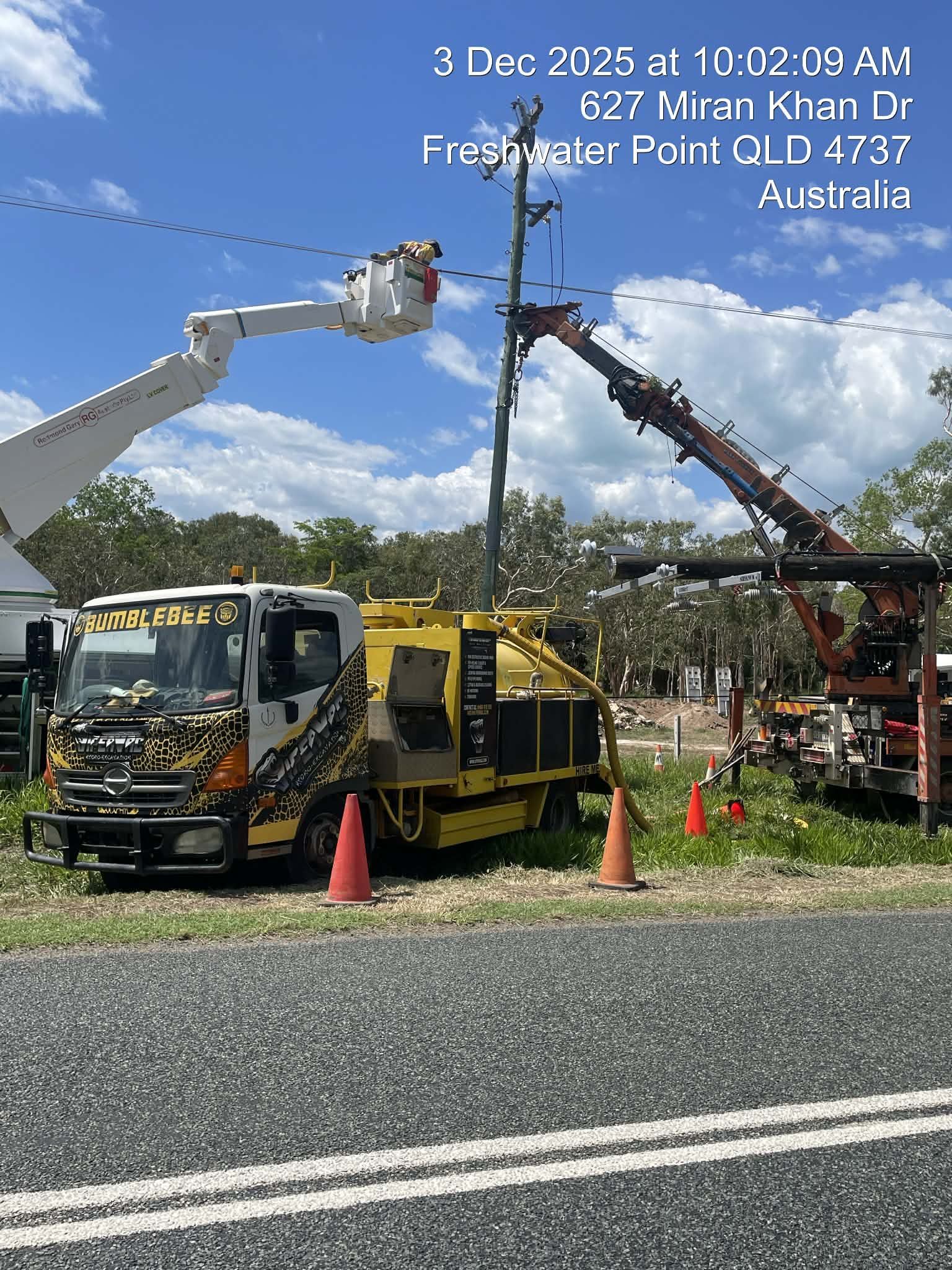 Utility trucks and boom lifts working on roadside power lines in a rural area under a cloudy sky.