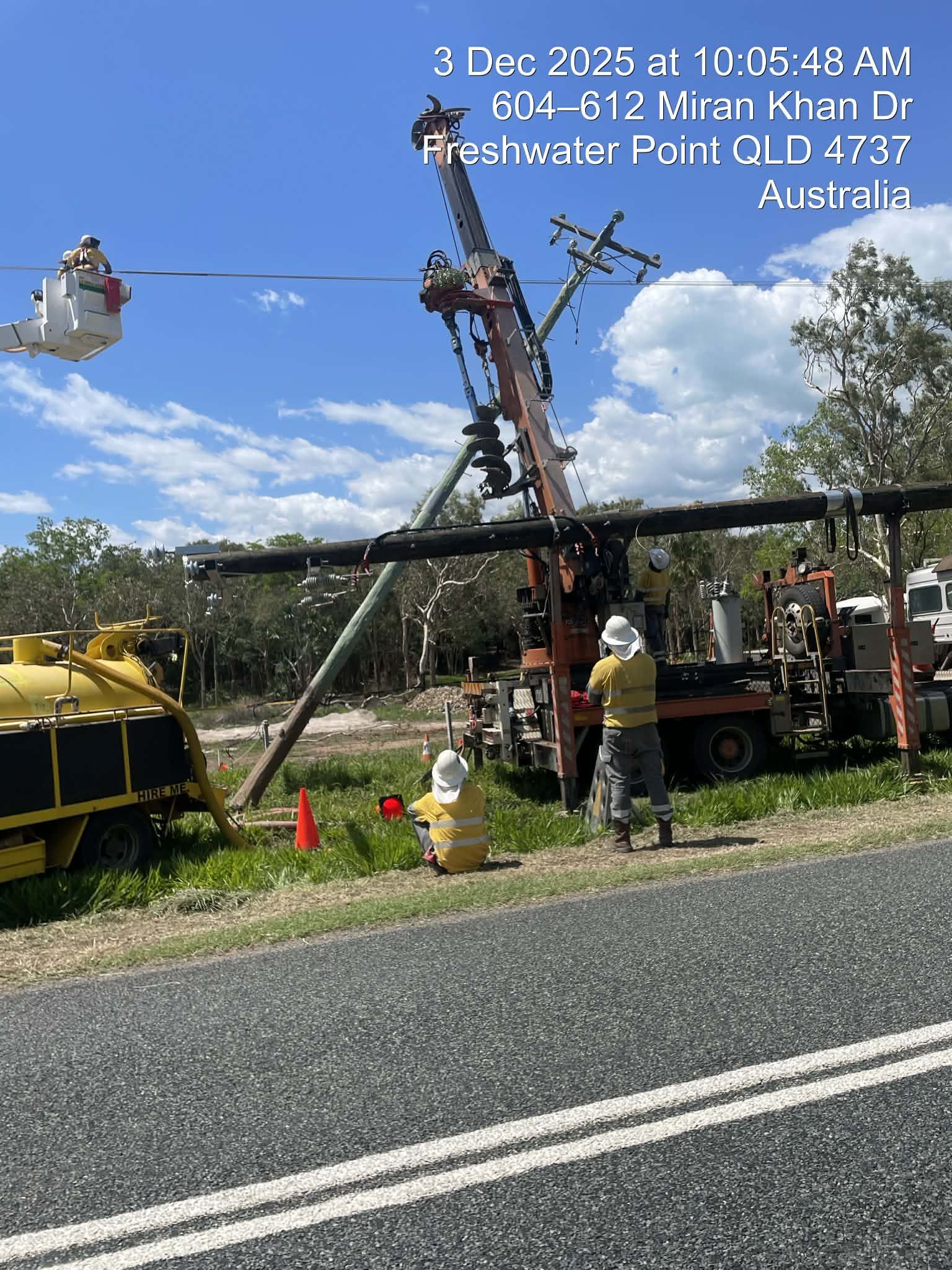 Utility workers repair a downed power pole beside a road, with a yellow tank and truck in the background.