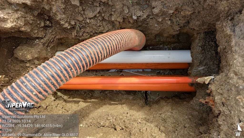 Orange corrugated conduit and cables in a dirt trench beside gray and orange pipes under a wall opening