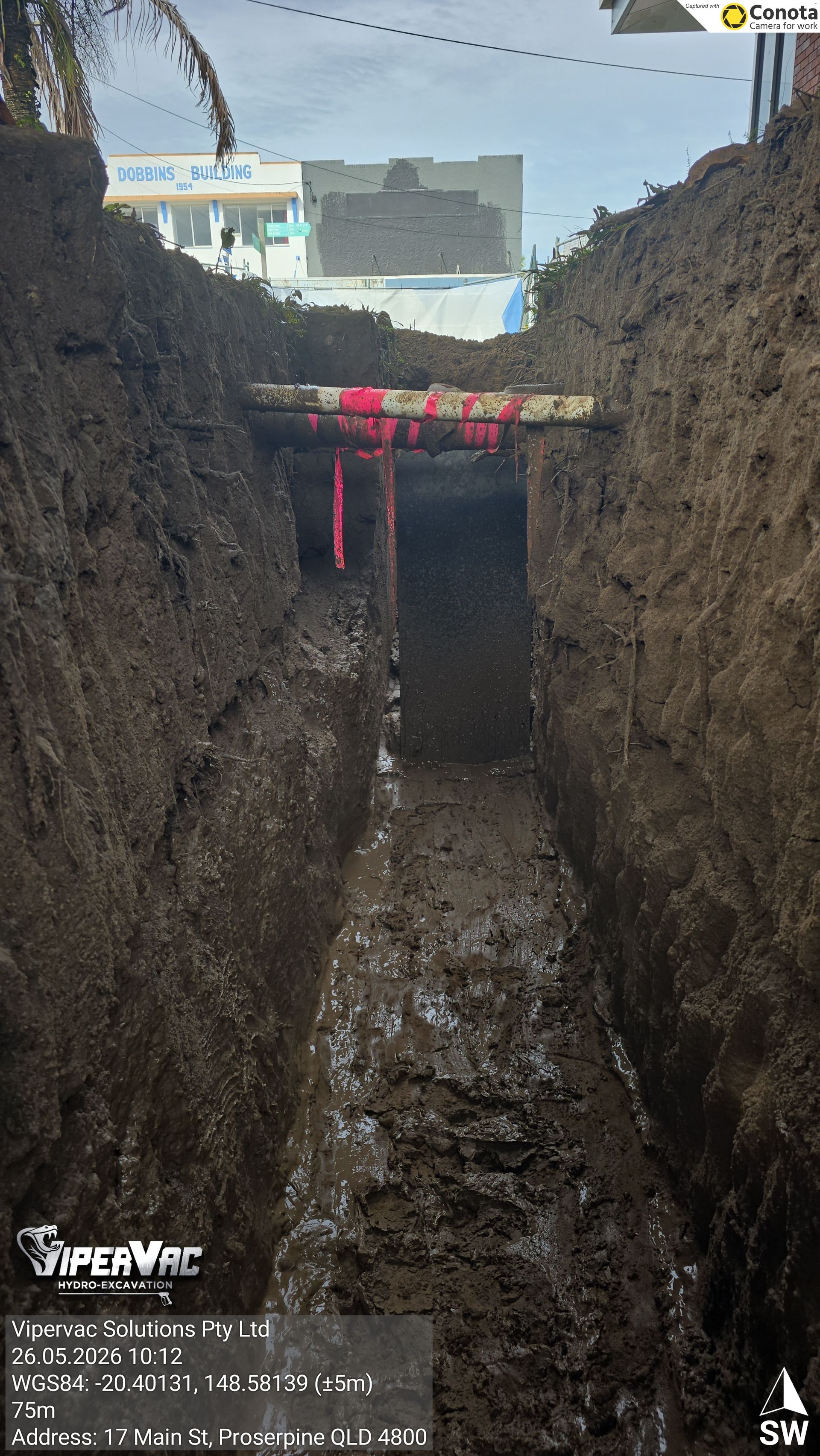 Deep trench with muddy floor and vertical earthen walls, red barrier tape across the top under a bright sky.