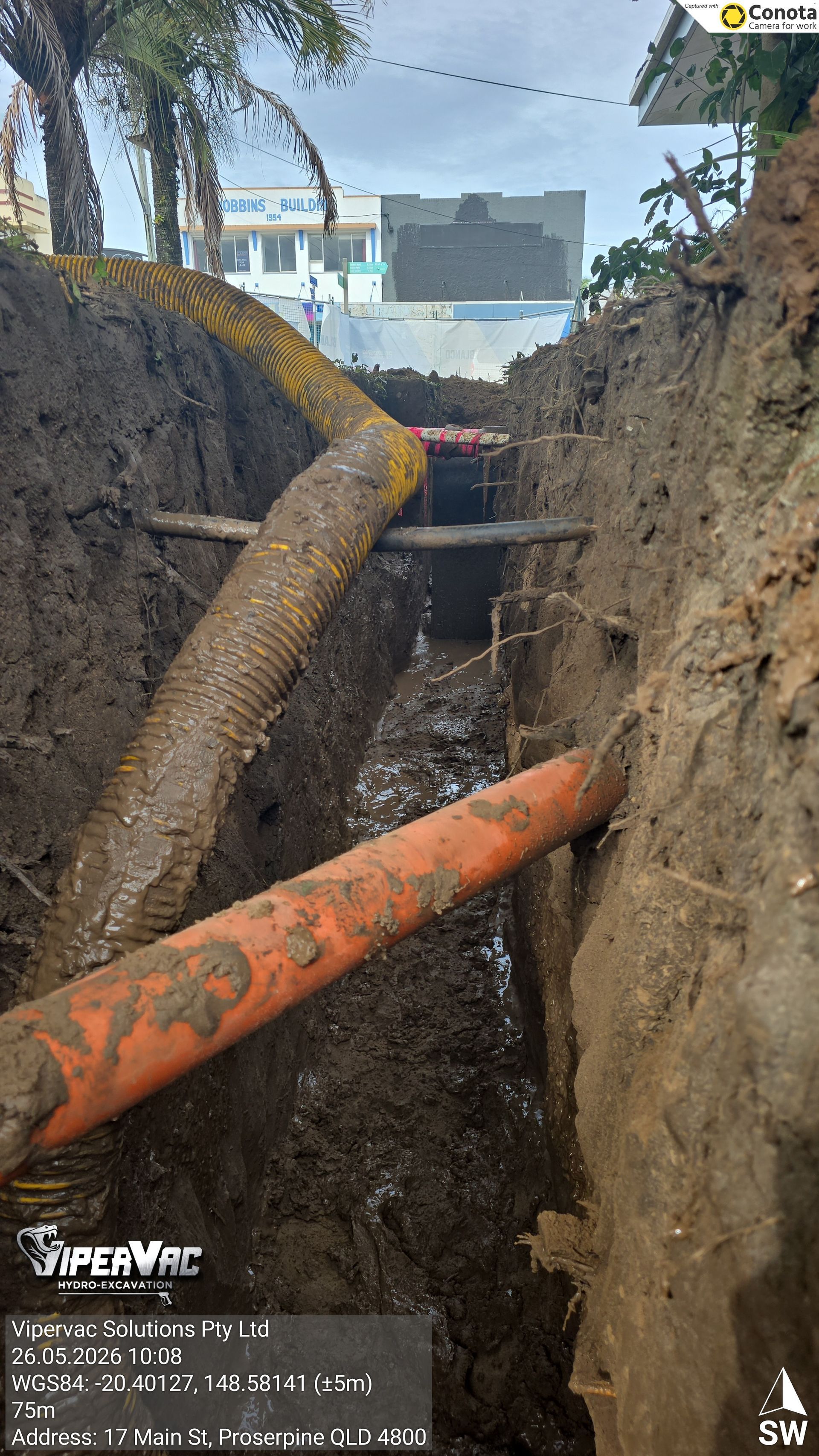 Excavation trench with two large pipes crossing muddy soil, construction site in the background