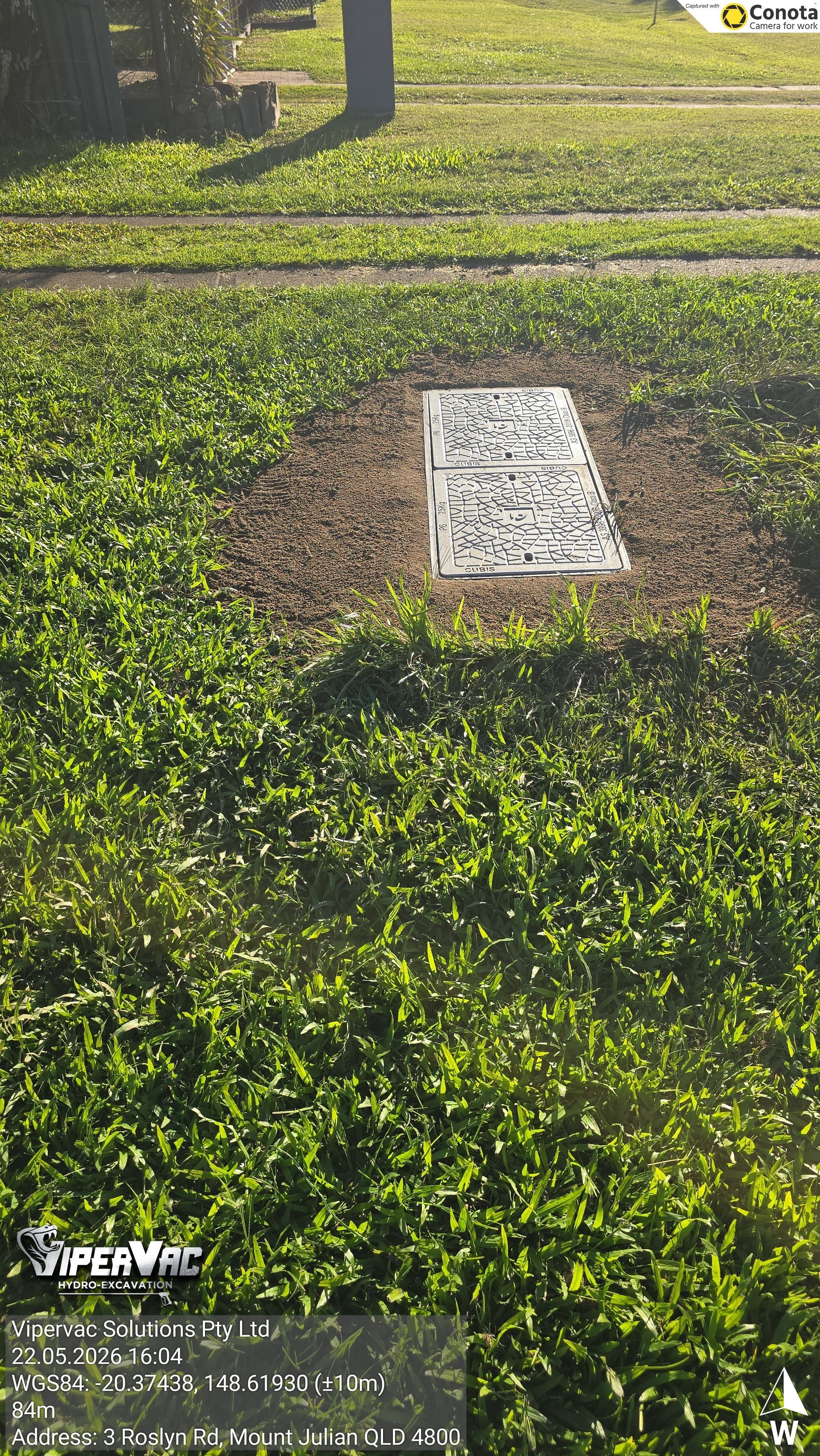 White rectangular utility marker in green grass near a paved walkway, with a tree trunk at left.
