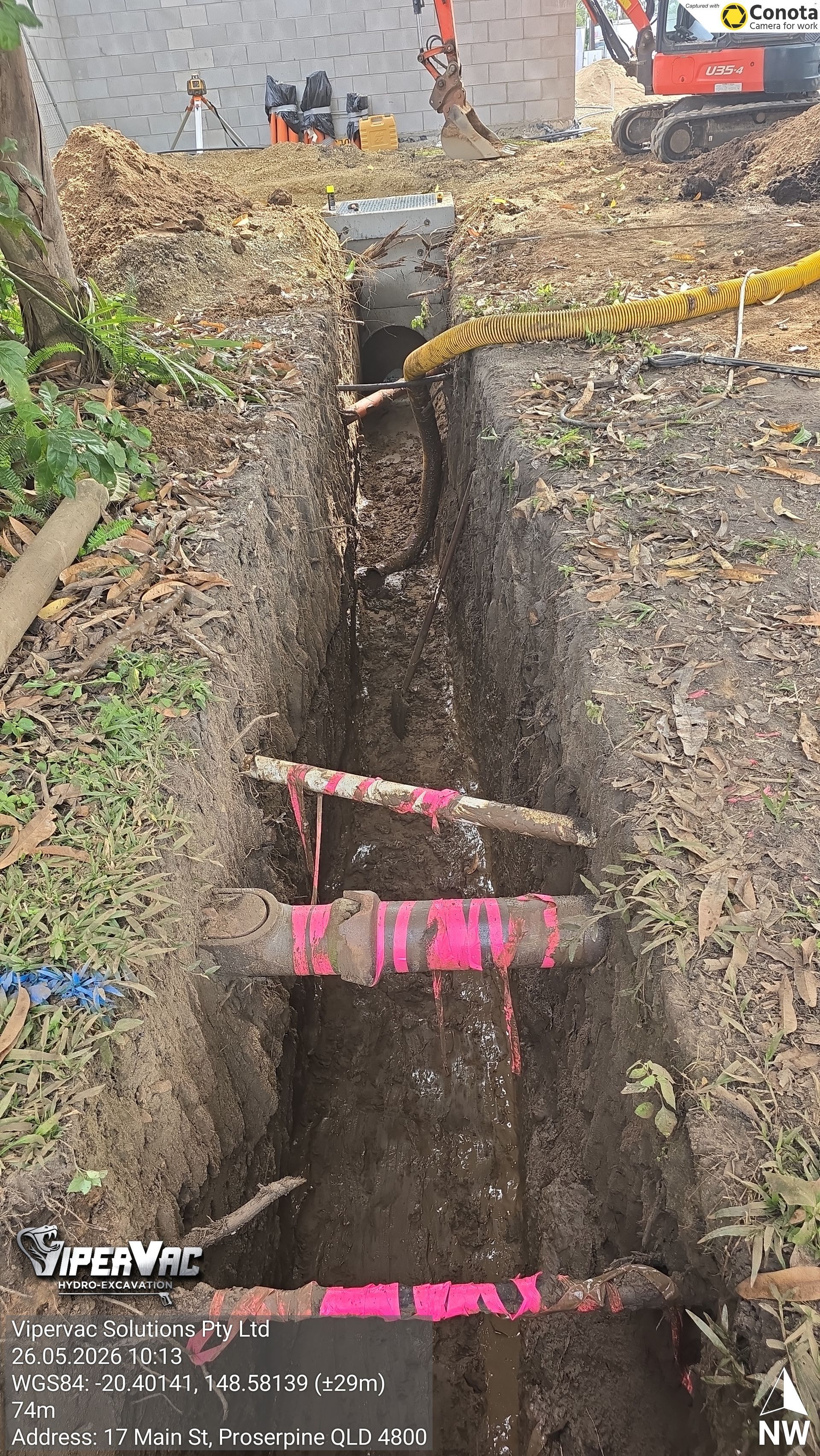 Narrow muddy trench beside a path, marked with pink tape and yellow caution tape.