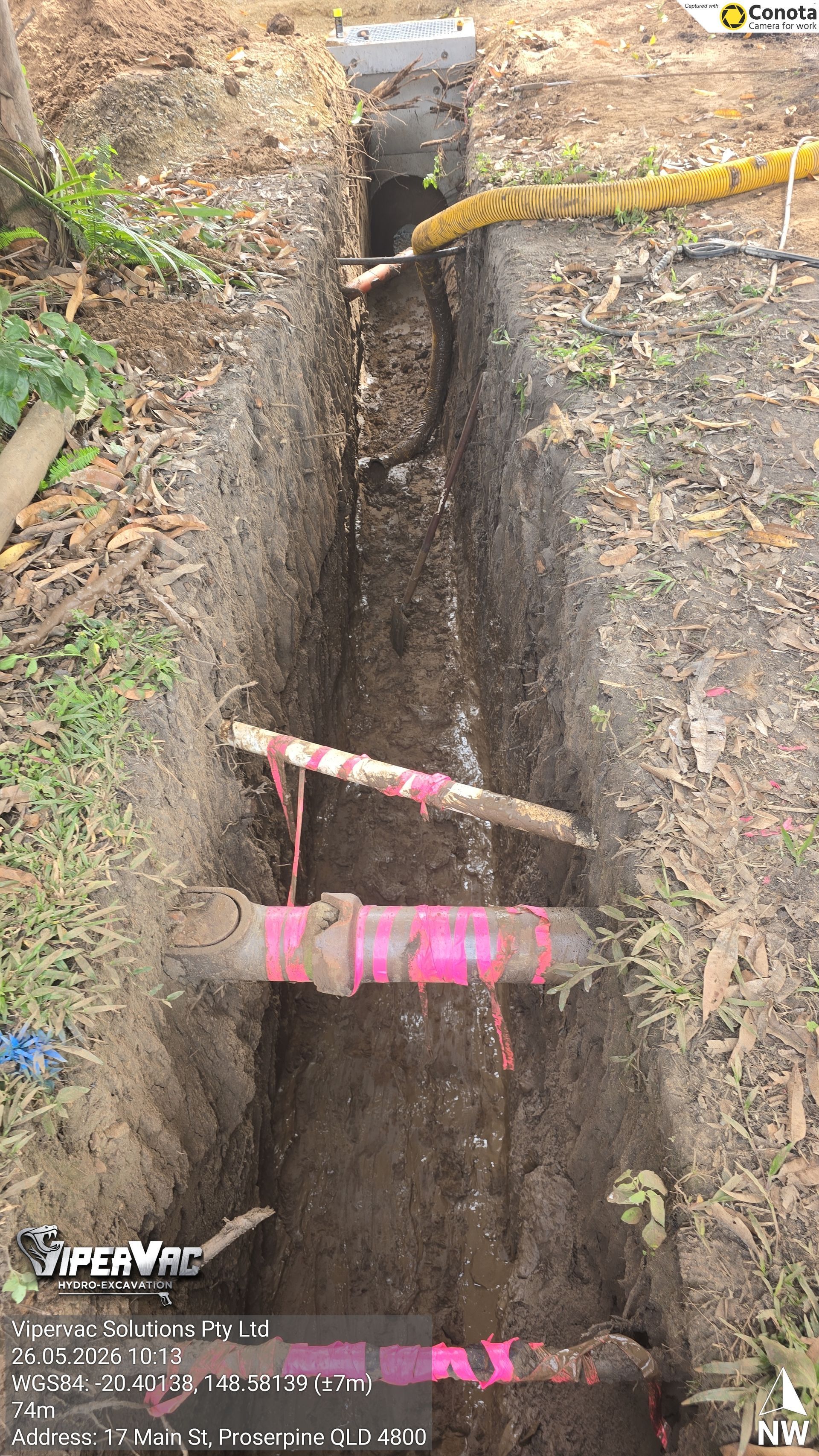 Deep trench with exposed utility pipes and a yellow hose in muddy ground; a brick utility box stands at the top.