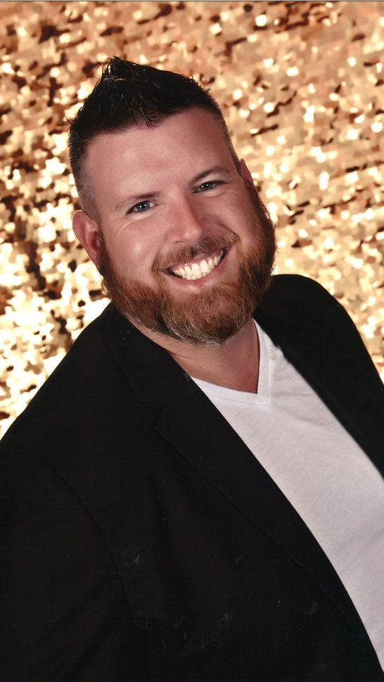 A person with a beard smiling, wearing a white shirt and black blazer against a gold sequined backdrop.