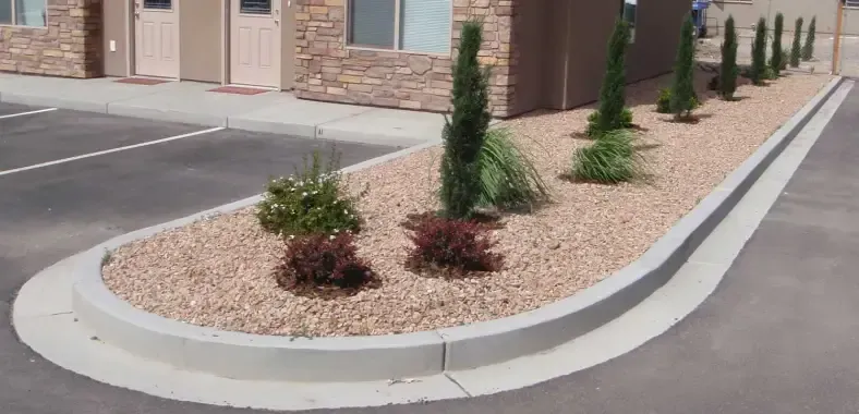 Curved flower bed with light brown gravel, shrubs, and trees along a building's edge; a gray curb separates it from the pavement.