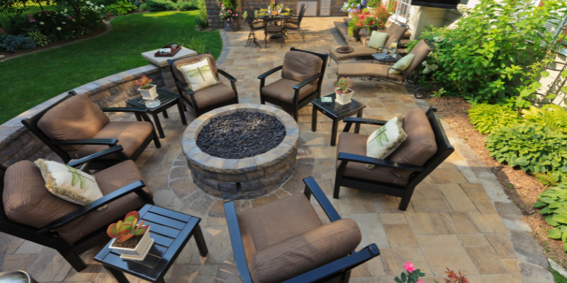 Patio with fire pit, seating, and green landscaping. Brown chairs surround a stone fire pit.