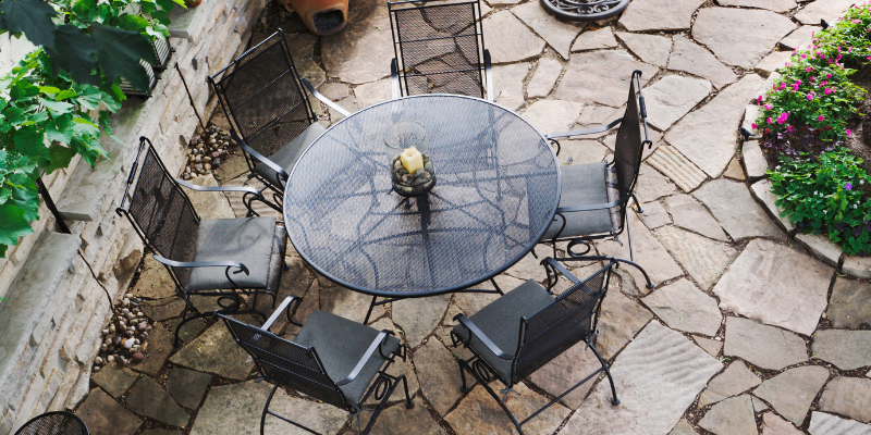 Overhead view of a patio with a wrought iron table and chairs on a stone surface. Green plants and flowers are visible.