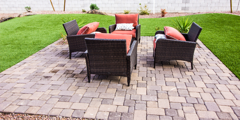 Patio with four wicker chairs, orange cushions and pillows, on a brick paved surface, surrounded by grass.