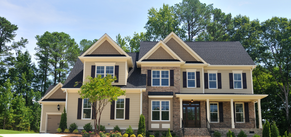 Two-story house with tan siding, brick facade, black shutters, and dark roof, set against a backdrop of trees and a blue sky.