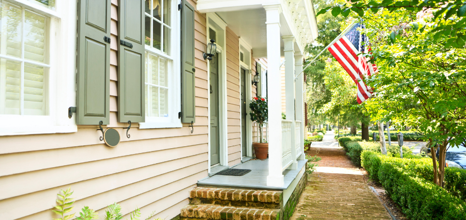 A residential house with a porch and American flag. A brick pathway and hedges in the front.