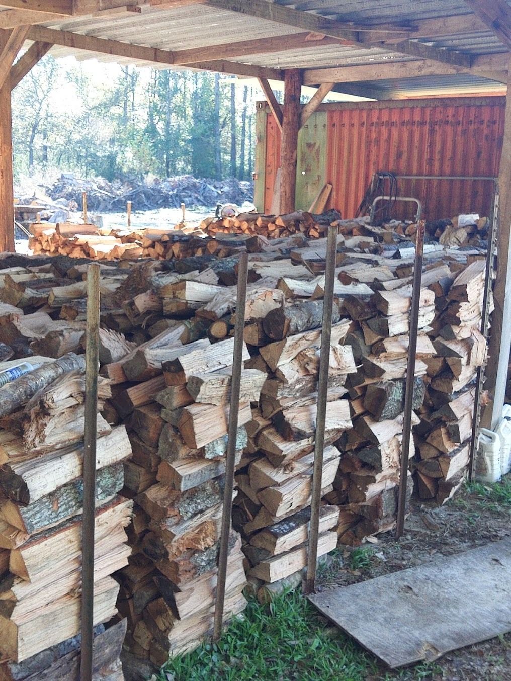 Stacked firewood under a rustic wooden shelter with a red shed in the background.