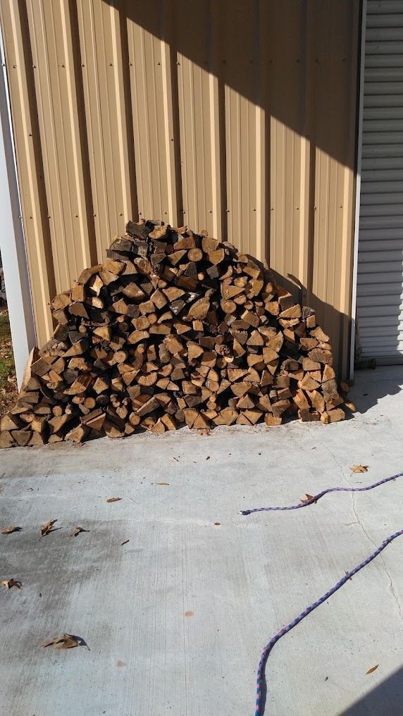 Stack of split firewood against a beige wall on a concrete patio, with a blue cord nearby.
