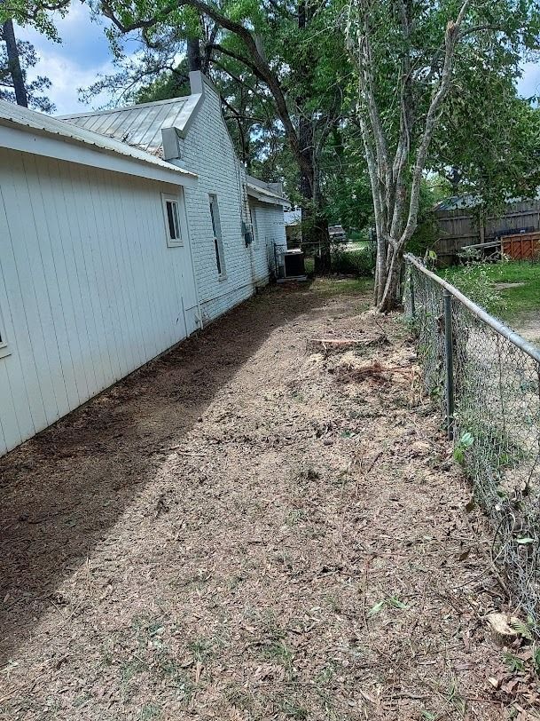 Narrow dirt path beside a white house and chain-link fence, lined with trees and a shaded yard.
