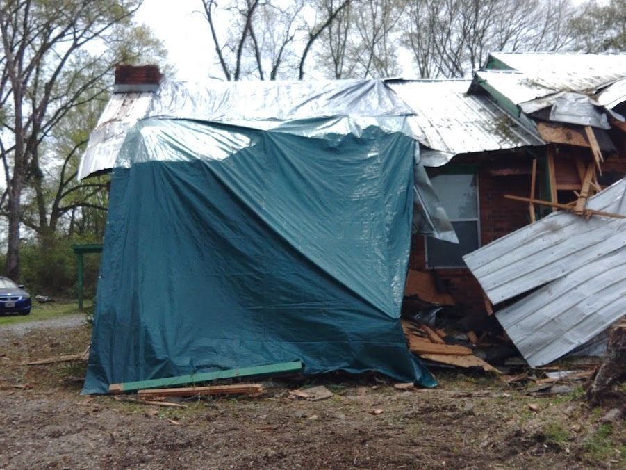 Damaged house with tarp-covered wall and collapsed metal roofing in a wooded yard