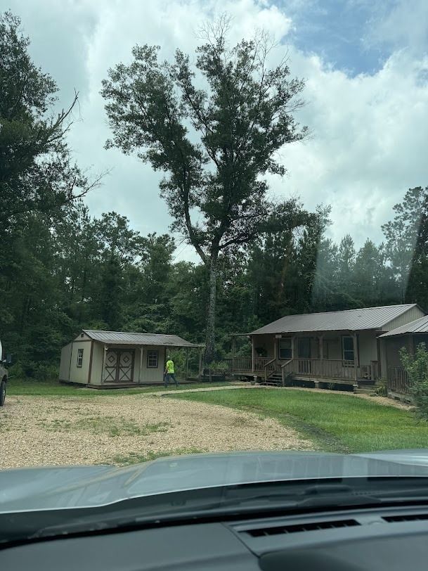 Rural campground with small cabins, gravel driveway, and tall trees under a cloudy sky.