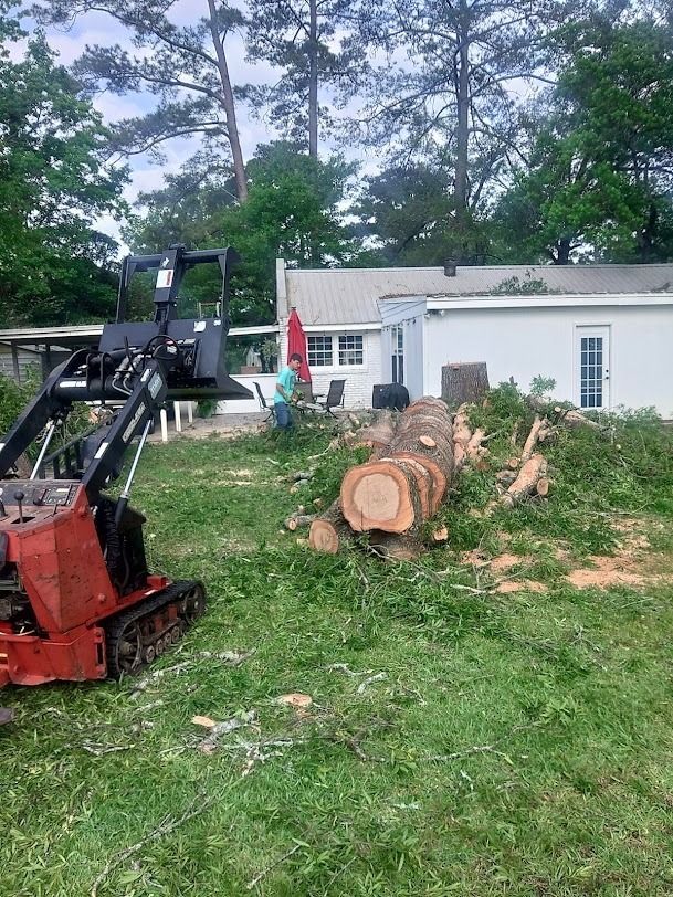 Red skid-steer near cut logs beside a white house in a grassy yard