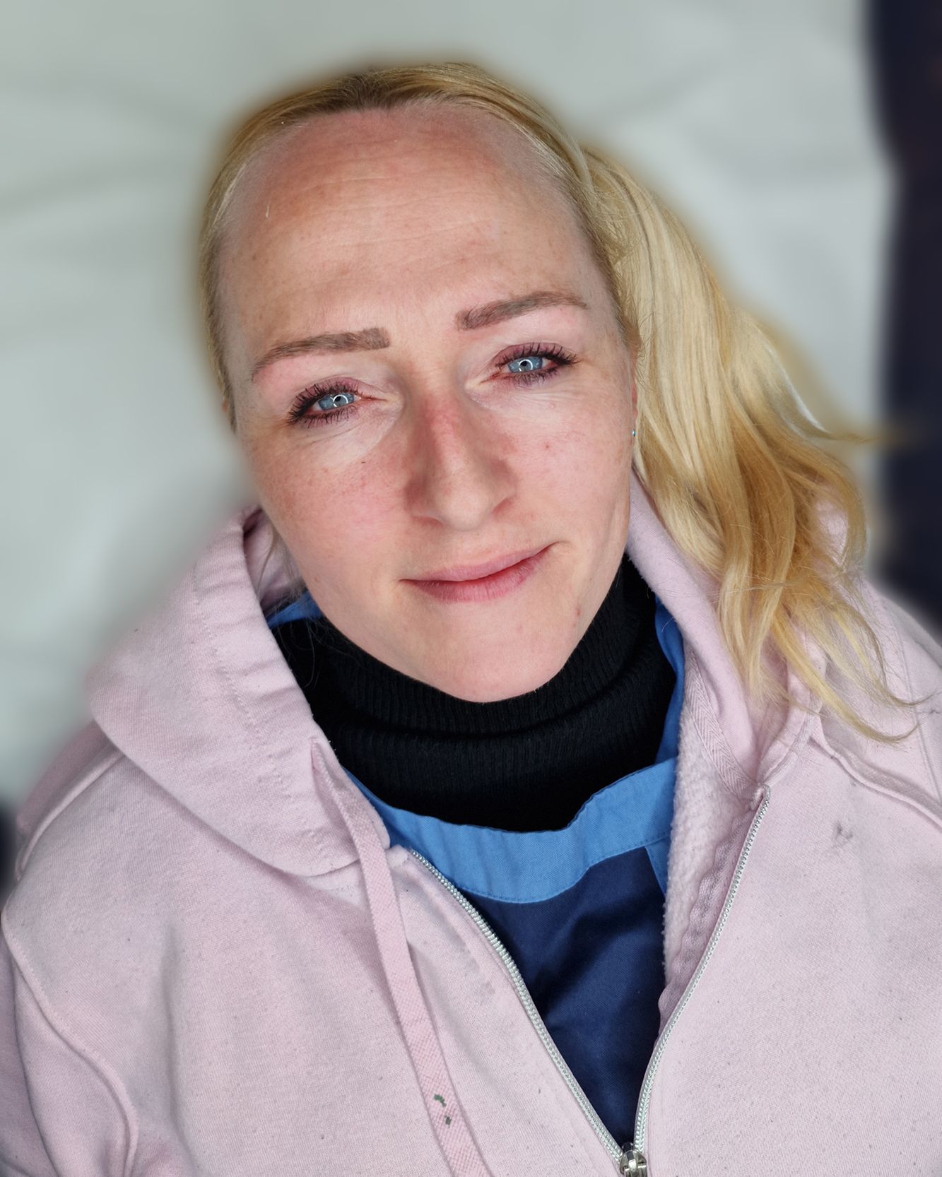 Blond-haired person wearing a pink hoodie and black turtleneck; looking at the camera.