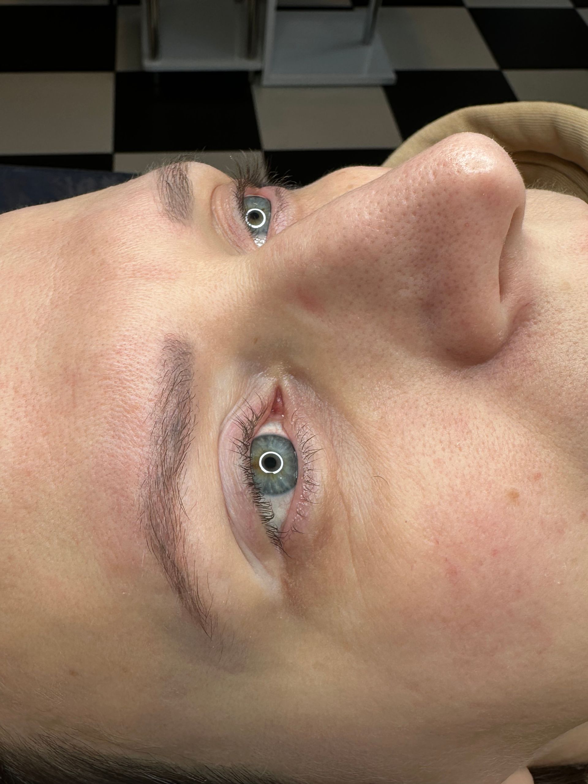 Close-up of a person's face with new eyebrow and eyeliner tattoos. Pale skin, blue eyes, checkerboard floor.