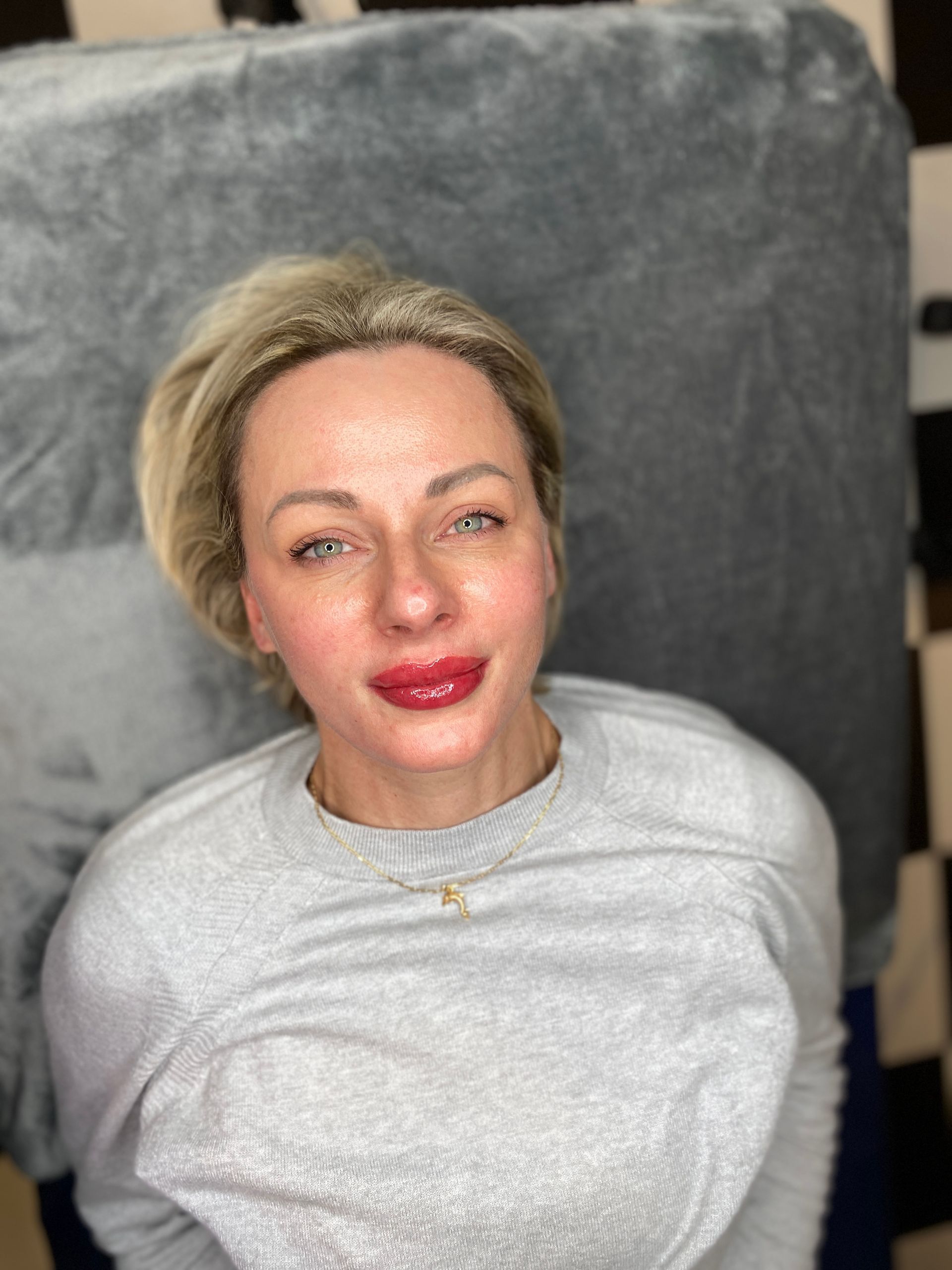 Woman with blonde hair and red lips, wearing a gray sweater, lying on a gray surface, looking up.