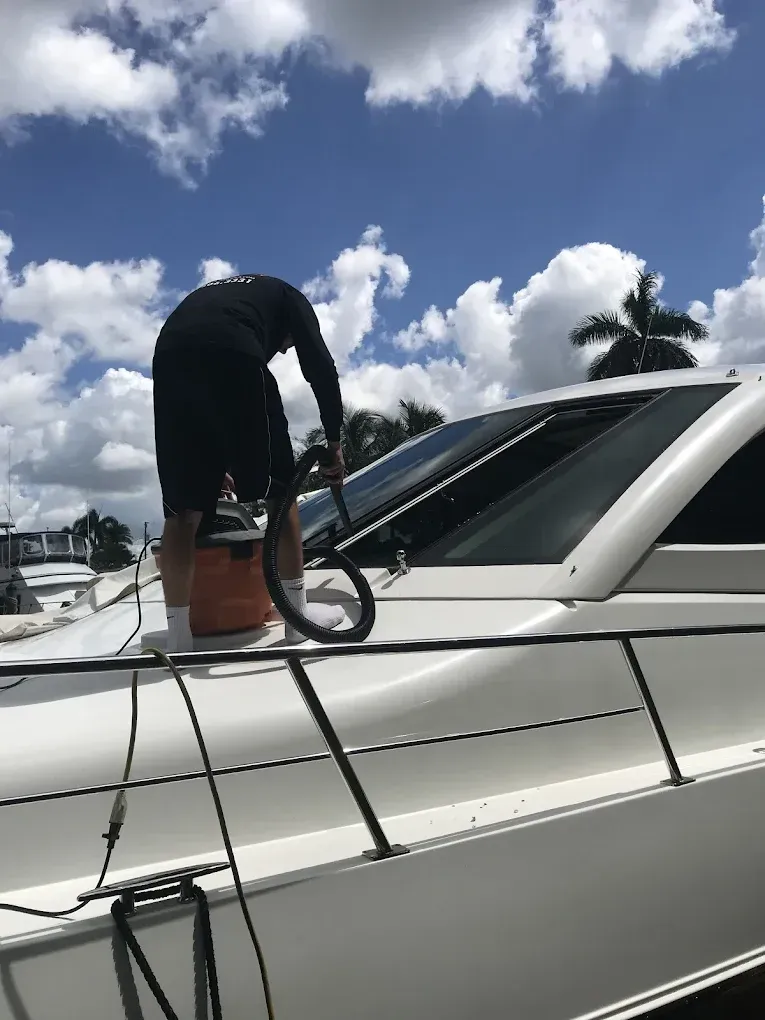 Person vacuuming a white boat deck with blue sky and clouds in the background.