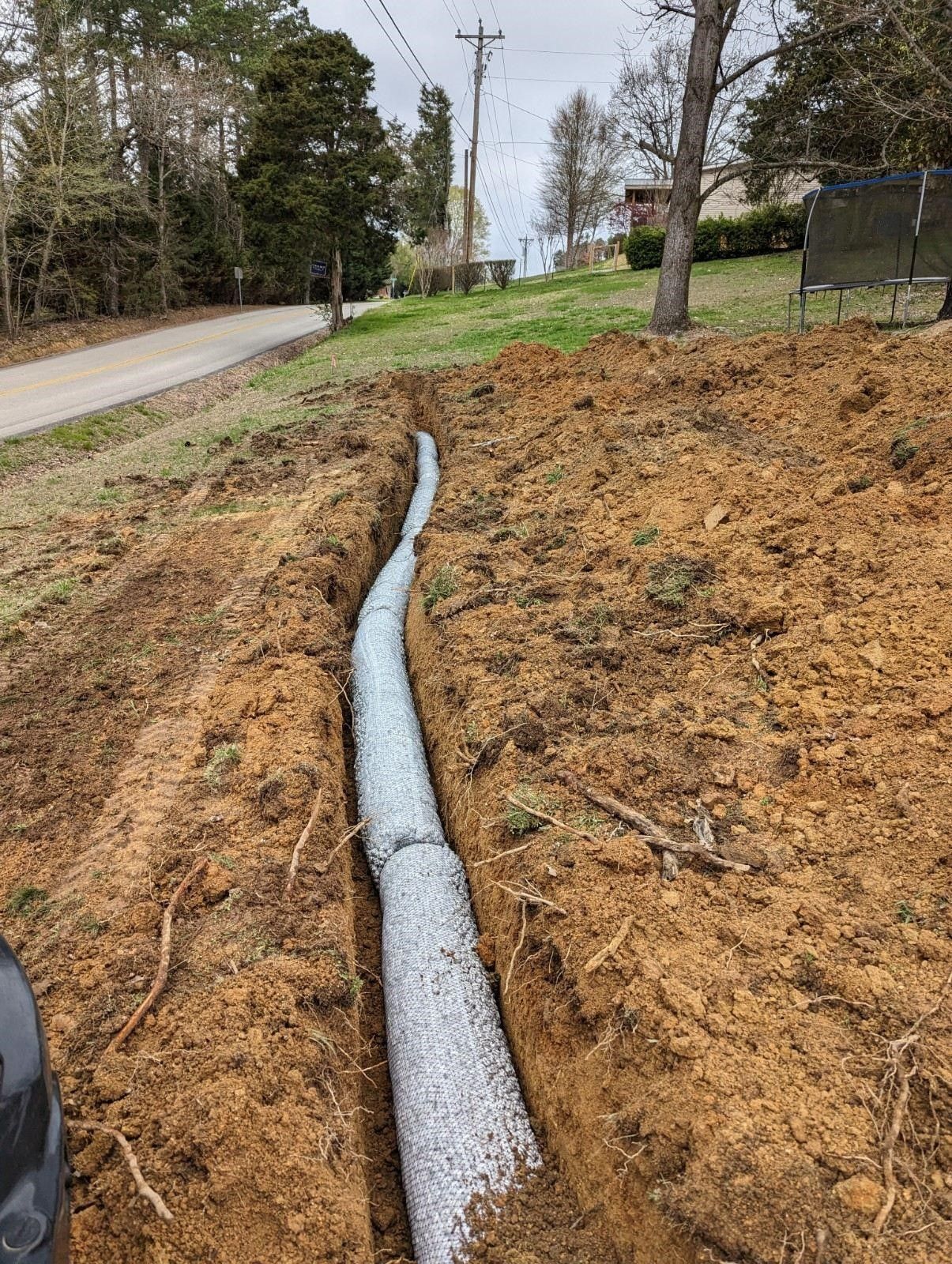 Black pipe in a brown dirt slope beside a road and grassy area under cloudy skies.