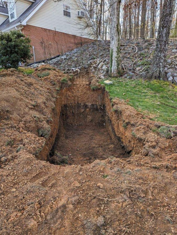 Deep rectangular trench dug in a backyard beside a house, with piles of excavated soil around it.
