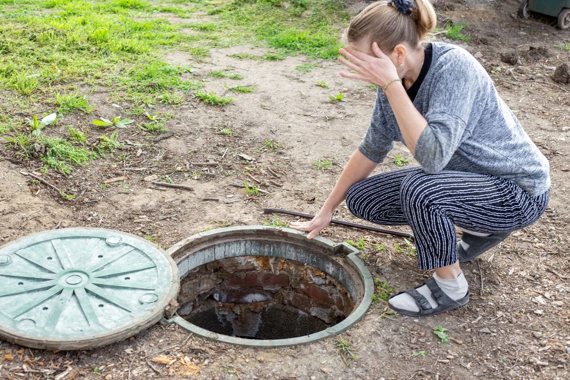 Woman crouching beside an open manhole on a dirt path, covering her face with one hand.