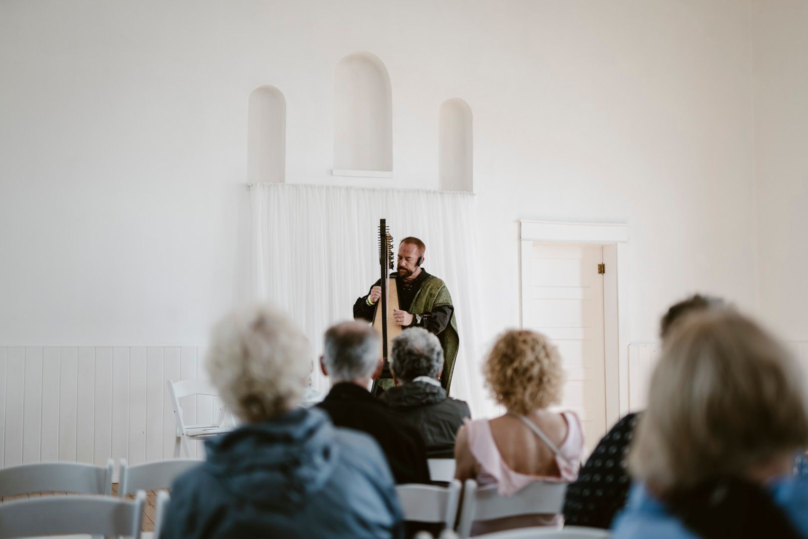 A musician performs on a string instrument before a small, seated audience in a bright room with arched wall alcoves.