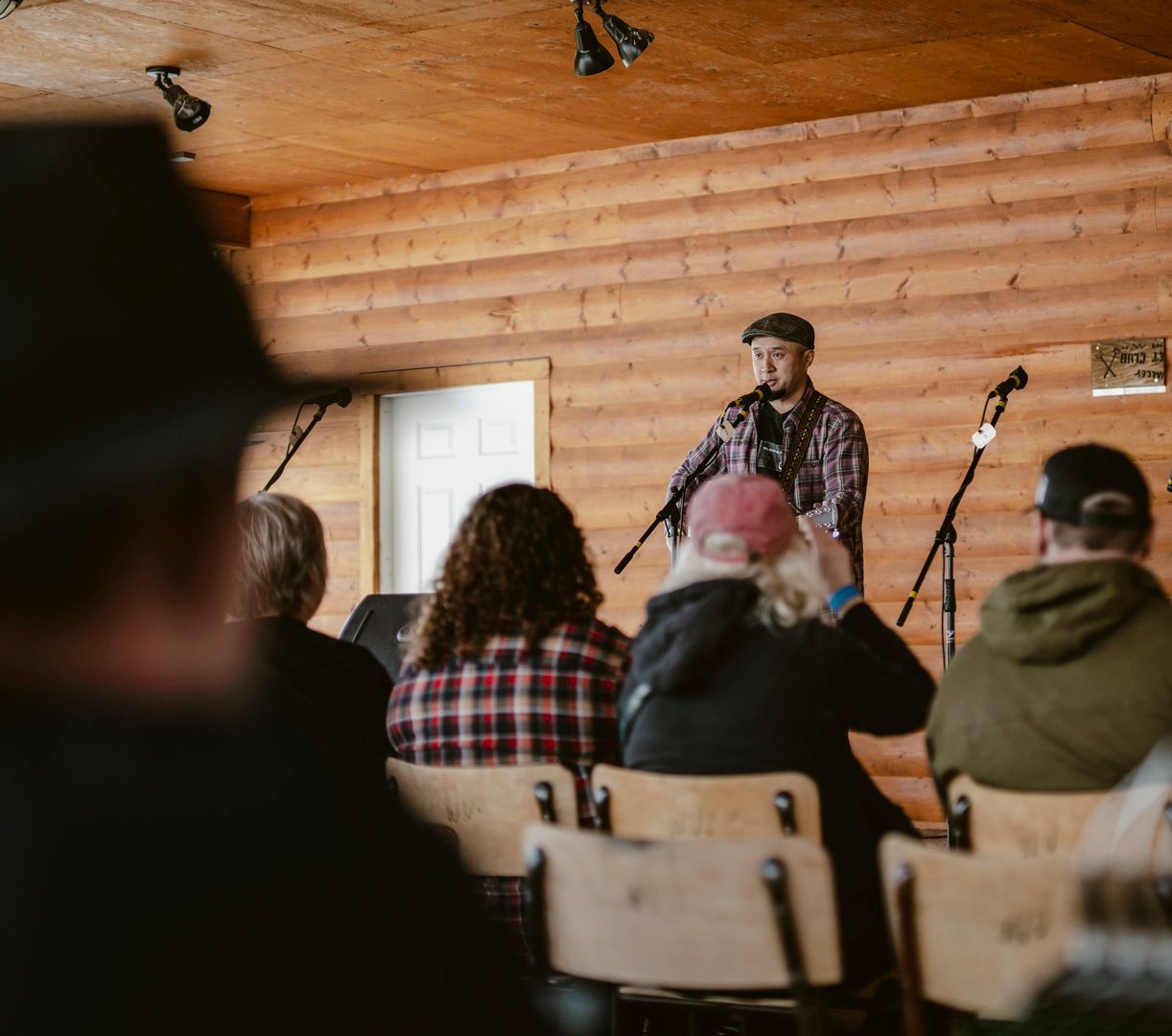 A musician performs at a microphone in a log-walled room, seen from behind an audience seated in rows of wooden chairs.